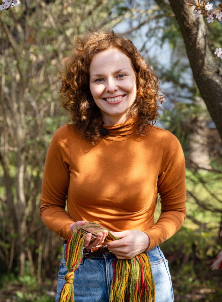 Young woman in orange blouse, curly hair, holding a set of weaving tablets and threads; tree in the background.